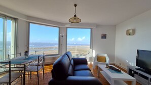 Una sala moderna con un sofá grande, una mesa de comedor y un televisor. La habitación tiene ventanas amplias con vista a la playa y al océano.