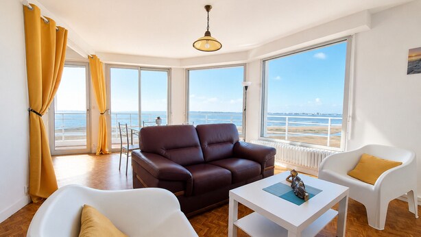 Un salón moderno con un sofá de cuero marrón, sillones blancos y una mesa de centro blanca. Grandes ventanales ofrecen vistas a la playa.