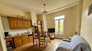 A spacious room with wooden flooring, a dining area with wooden chairs and a table, a bed with a grey blanket, and a large window providing natural light. - Family apartment for 4 in the heart of La Bourboule (La Bourboule)