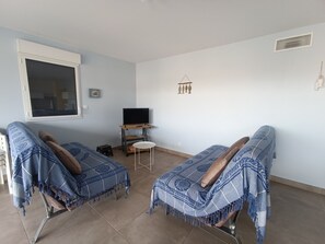 Habitació amb dos sofàs coberts amb mantilles blaves amb dibuixos, una taula petita amb un televisor sobre un suport i una finestra amb persianes.