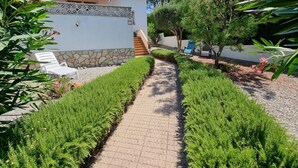 Un sentiero conduce a un edificio con base in pietra e tetto rosso, circondato da siepi curate e alberi.