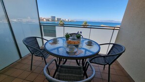Une table ronde en verre, entourée de quatre chaises en osier, sur un balcon donnant sur un plan d’eau et des bâtiments.