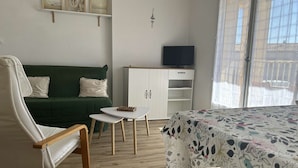 Ein Zimmer mit einem grünen Sofa, einem weißen Sessel, einem kleinen weißen Tisch, einem Bett mit floralem Überwurf und einem Fernseher auf einem Schrank. Ein Fenster mit Vorhängen ist ebenfalls zu sehen.