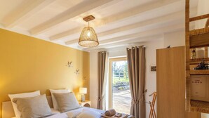 A bedroom with a bed, bedside lamp, curtains, and a view of the outdoors. - Cozy countryside cottage, 2 bedrooms, open views (Baugé en Anjou)