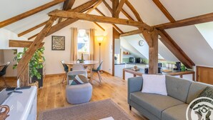 An attic-style room with exposed wooden beams, a dining area with a table and chairs, a sofa, and a kitchen area with a microwave. - Cozy countryside cottage, 2 bedrooms, open views (Baugé en Anjou)