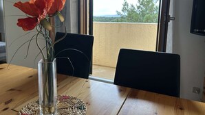 Ein Esstisch mit einem Blumenstrauß in einer Vase, einer gewebten Tischmatte und Blick auf einen Balkon mit Holzgeländer und einer Tür.