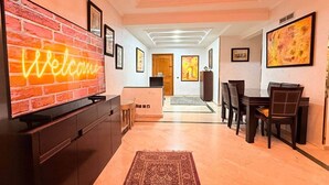 Ein modernes Wohnzimmer mit Fernseher, der auf einer Ziegelwand 'Welcome' anzeigt, ein Essbereich mit Tisch und Stühlen sowie ein Flur, der zu einer Tür führt.