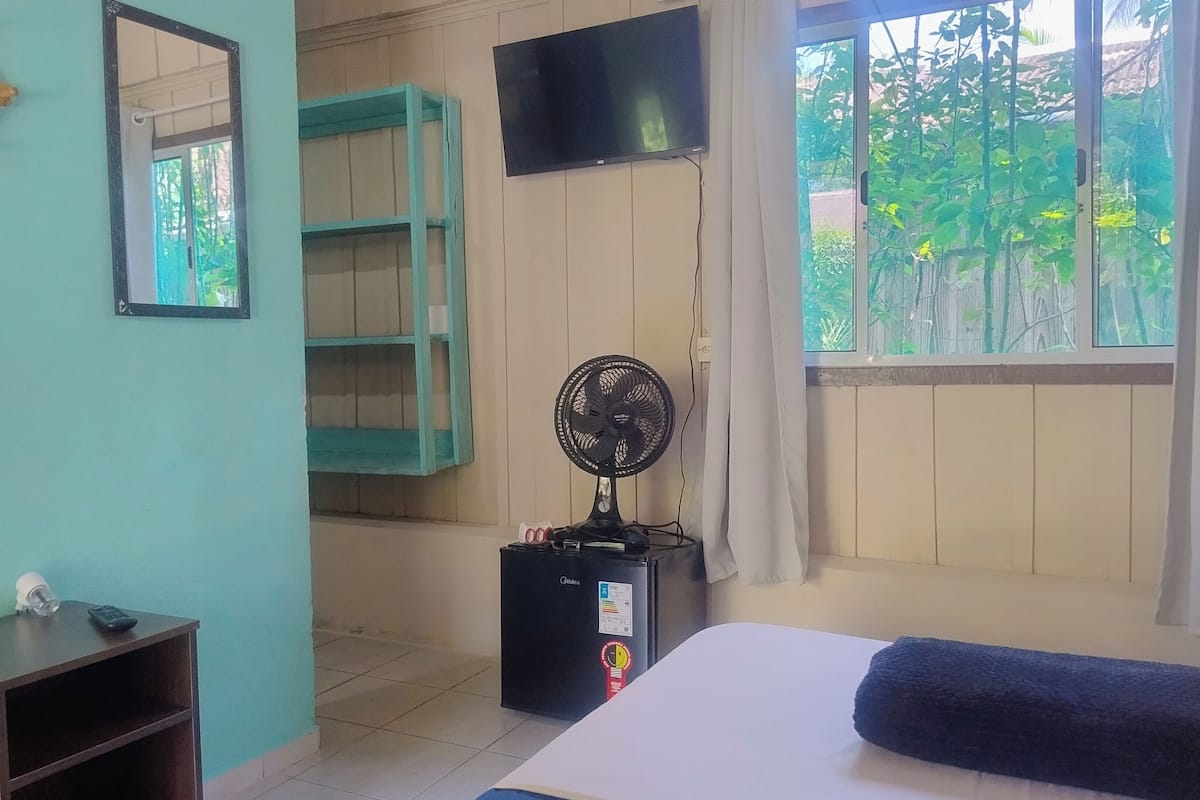 A room with a bed, a standing fan, a TV mounted on the wall, and a window with a view of greenery.