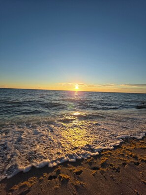 Ein Strand mit sanft anschwellenden Wellen und einer untergehenden Sonne in der Ferne.