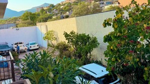 Un patio con un coche blanco estacionado cerca de un edificio, rodeado de follaje y una montaña al fondo.