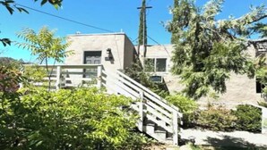 Un edificio residencial con una escalera que conduce a una puerta, rodeado de vegetación y un cielo despejado.