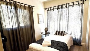 Un dormitorio con cama, cortinas, una mesita pequeña y un cuadro colgado en la pared.