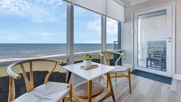 Un balcon avec vue sur l’océan, des chaises en bois, une petite table avec une plante, et une porte donnant sur un autre balcon équipé d’une chaise et d’une table.