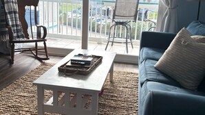 Ein Wohnzimmer mit einer Couch, zwei Sesseln, einem Couchtisch und Blick auf einen Balkon mit Geländer und Palmen.