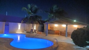 A swimming pool with a blue tint, a covered outdoor seating area, and palm trees.