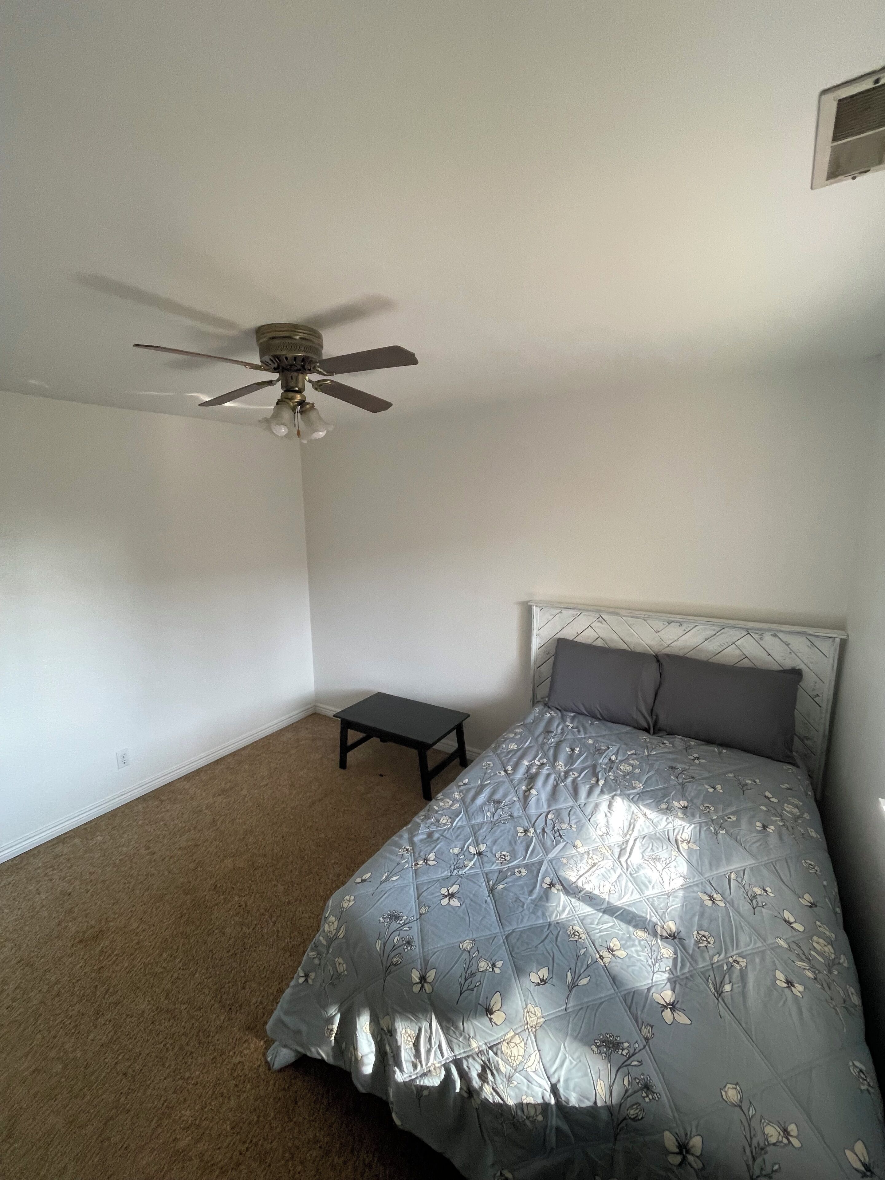 A bedroom with a single bed, a small table, a ceiling fan, and a plain white wall.
