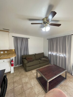 Ein Wohnzimmer mit Deckenventilator, einer Couch, einem Couchtisch und einer Küchenzeile inklusive Arbeitsplatte und Feuerlöscher.