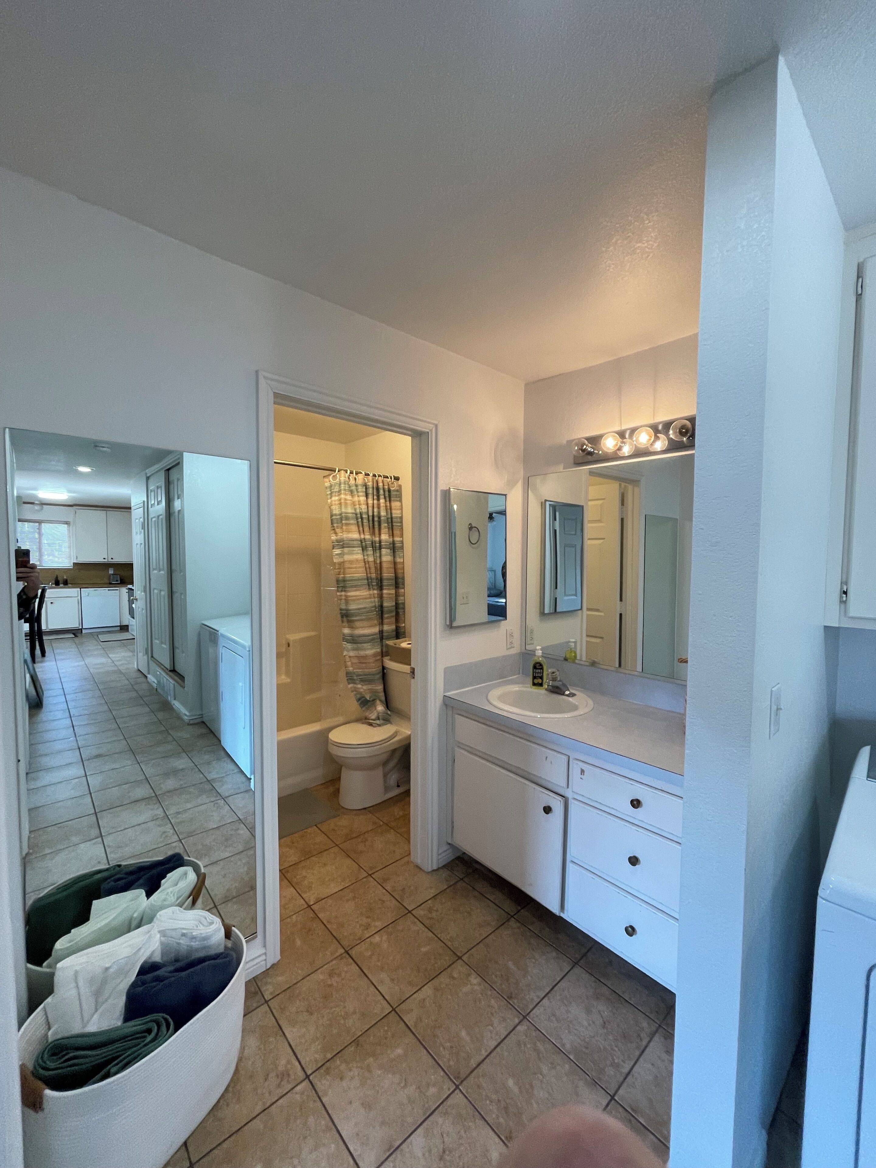 A bathroom with a toilet, sink, and mirror. There is a shower curtain and a storage bin with folded clothes.