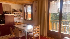 Una habitación acogedora con una mesa de comedor, sillas, un tapete rojo, piso de madera, una ventana con cortinas y vistas a un balcón.