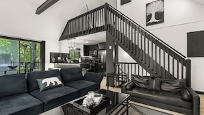 Una sala moderna con una escalera negra, un sofá negro con un cojín de oso, y un comedor con mesa de vidrio.