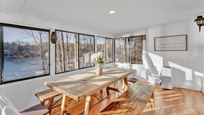 A dining area with a large wooden table and chairs, large windows with a view of trees and a body of water, and a wall-mounted lamp. - Lakefront: Hot Tub + Fire Pit + Game Room + Pool (Tobyhanna)
