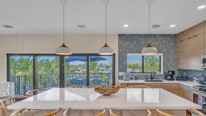 Cocina moderna con una isla grande, comedor con sillas, luces colgantes y vistas a palmeras a través de puertas de cristal.