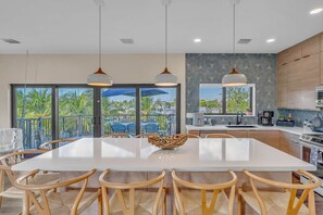 A modern kitchen with a large island, dining area with chairs, pendant lights, and a view of palm trees through the glass doors. - Waterfront Luxury, Volleyball, Putting Green, Dock & More! (Marathon)