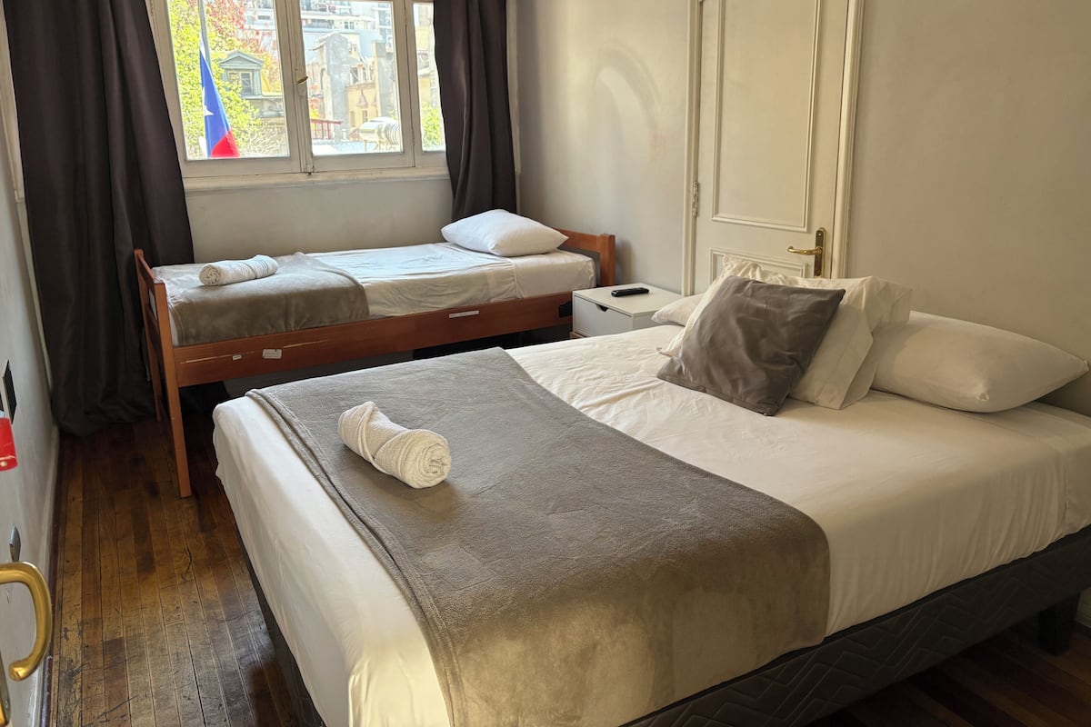 Een hotelkamer met twee enkele bedden, een houten vloer, een raam met gordijnen, een nachtkastje met lamp, en een deur met een deurklink.