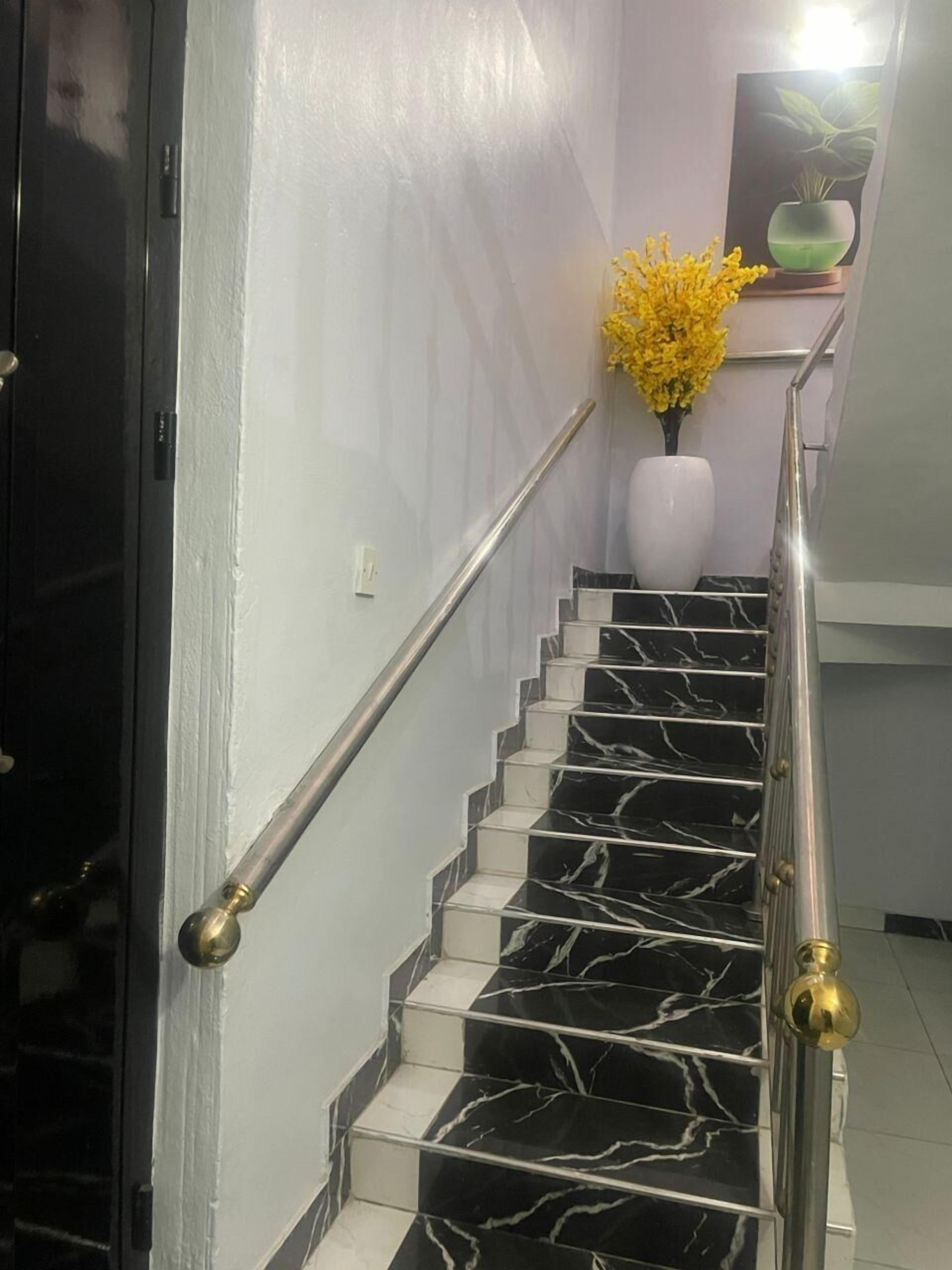 Una escalera con escalones de mármol blanco y negro, barandilla de metal y un jarrón blanco con flores amarillas en la parte superior.