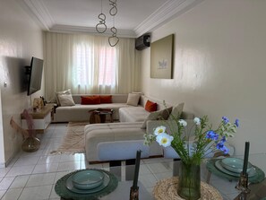 Ein Wohnzimmer mit einer Couch, einem Flachbildfernseher, einem Couchtisch und Dekorationselementen, darunter Blumen und Kerzen.