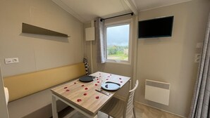 Ein kleiner Raum mit einem Holztisch für zwei Personen, einer Bank, einem an der Wand montierten Fernseher und einem Fenster mit Vorhängen.