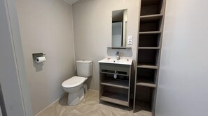 Un baño pequeño y moderno con inodoro, lavamanos y espejo. Hay un dispensador de papel higiénico en la pared y una estantería con varios compartimentos.