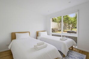 Quarto com duas camas, roupa de cama branca e uma grande janela com vista para um jardim.