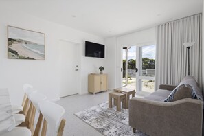 A modern living room with a flat-screen TV, a sofa, a dining area with chairs, and a view through the glass door. - Peaceful 2-Bed Apartment in Coastal McCrae (McCrae)