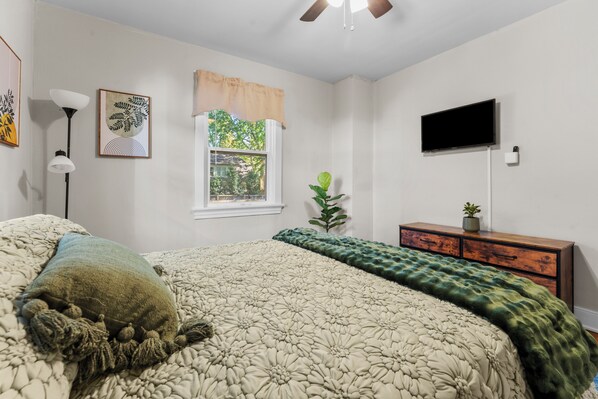 Une chambre à coucher comprenant un lit, une commode, un téléviseur et une fenêtre donnant sur un paysage verdoyant.