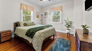 Ein Schlafzimmer mit einem Bett, einem Deckenventilator, zwei Fenstern mit Vorhängen, einem Nachttisch, einer Kommode und zwei Blumentöpfen.