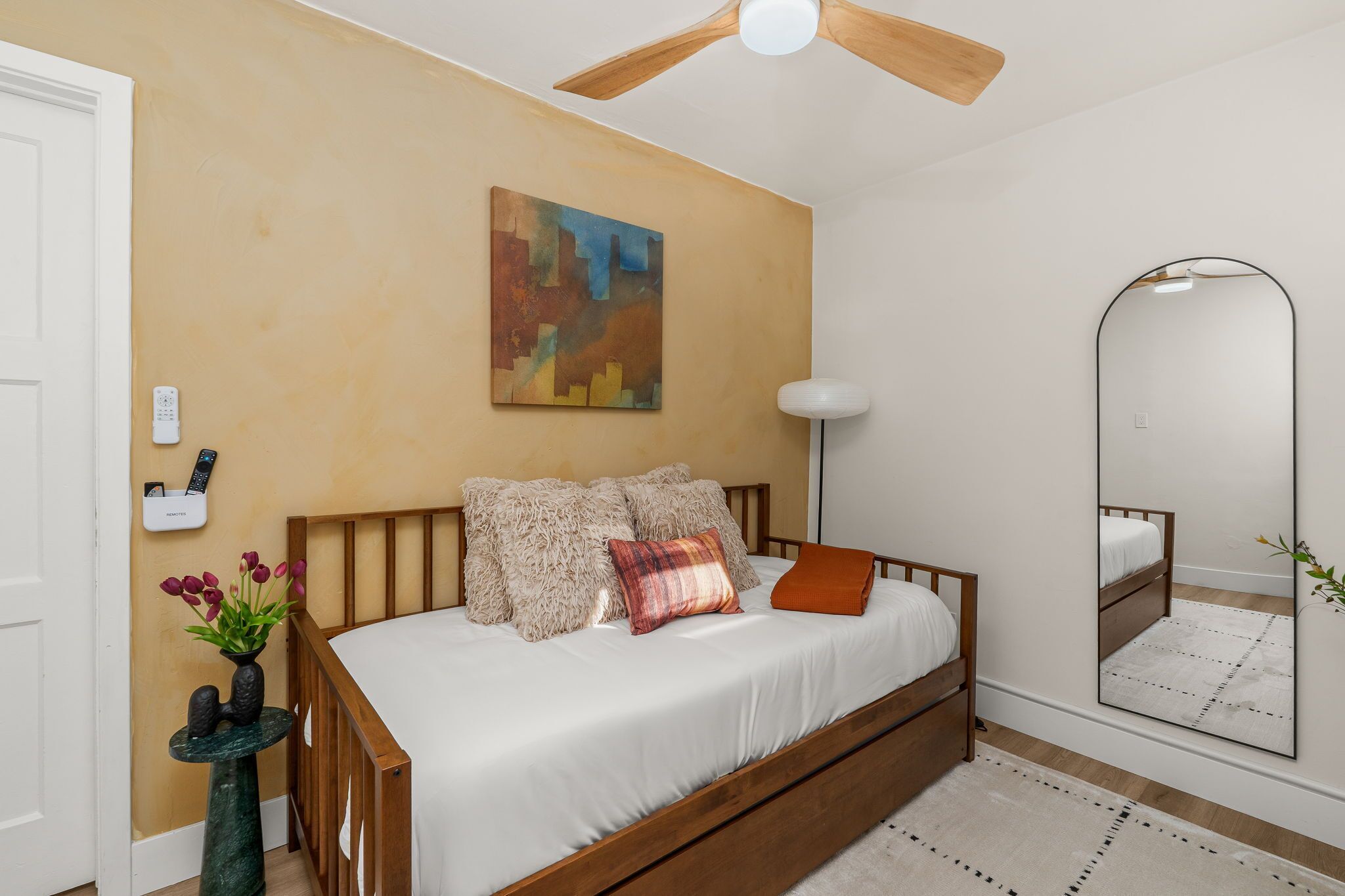 Een slaapkamer met een houten bed, een plafondventilator, een schilderij, een nachtkastje met een bloevas en een grote spiegel.