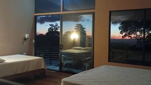 Ein Schlafzimmer mit zwei Betten, einem Balkon mit Sonnenuntergangsblick und einem großen Fenster.