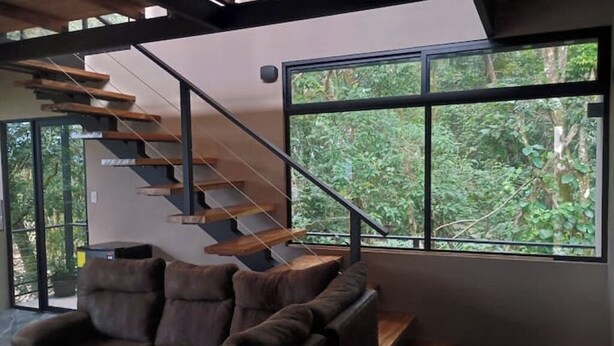 Ein modernes Interieur mit einer Holztreppe, einem braunen Ledersofa und großen Fenstern mit Blick auf Grünflächen.
