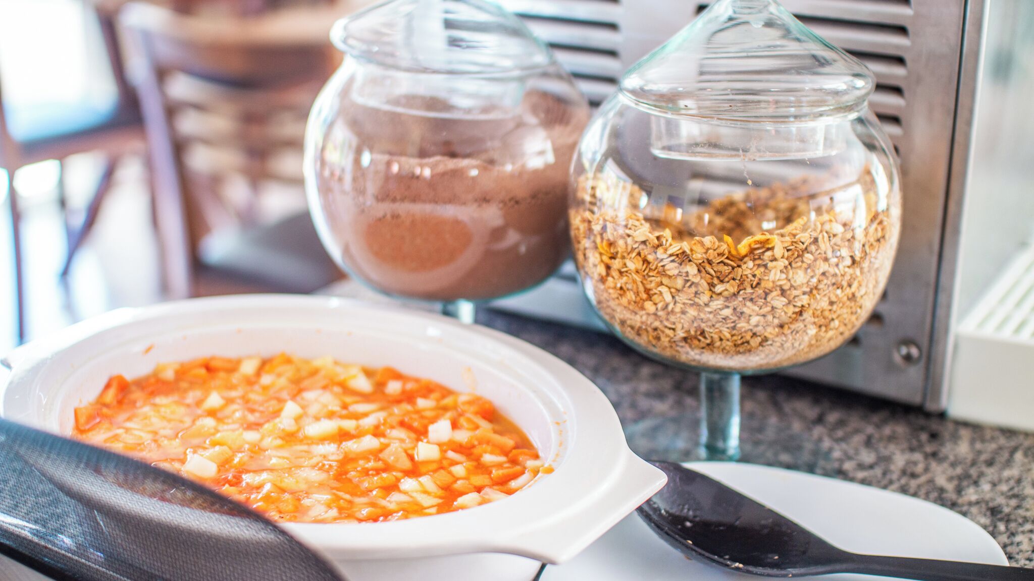 Un plan de travail de cuisine avec un saladier rempli de fruits coupés en dés, deux bocaux en verre contenant un liquide brun et des céréales, et une spatule noire.