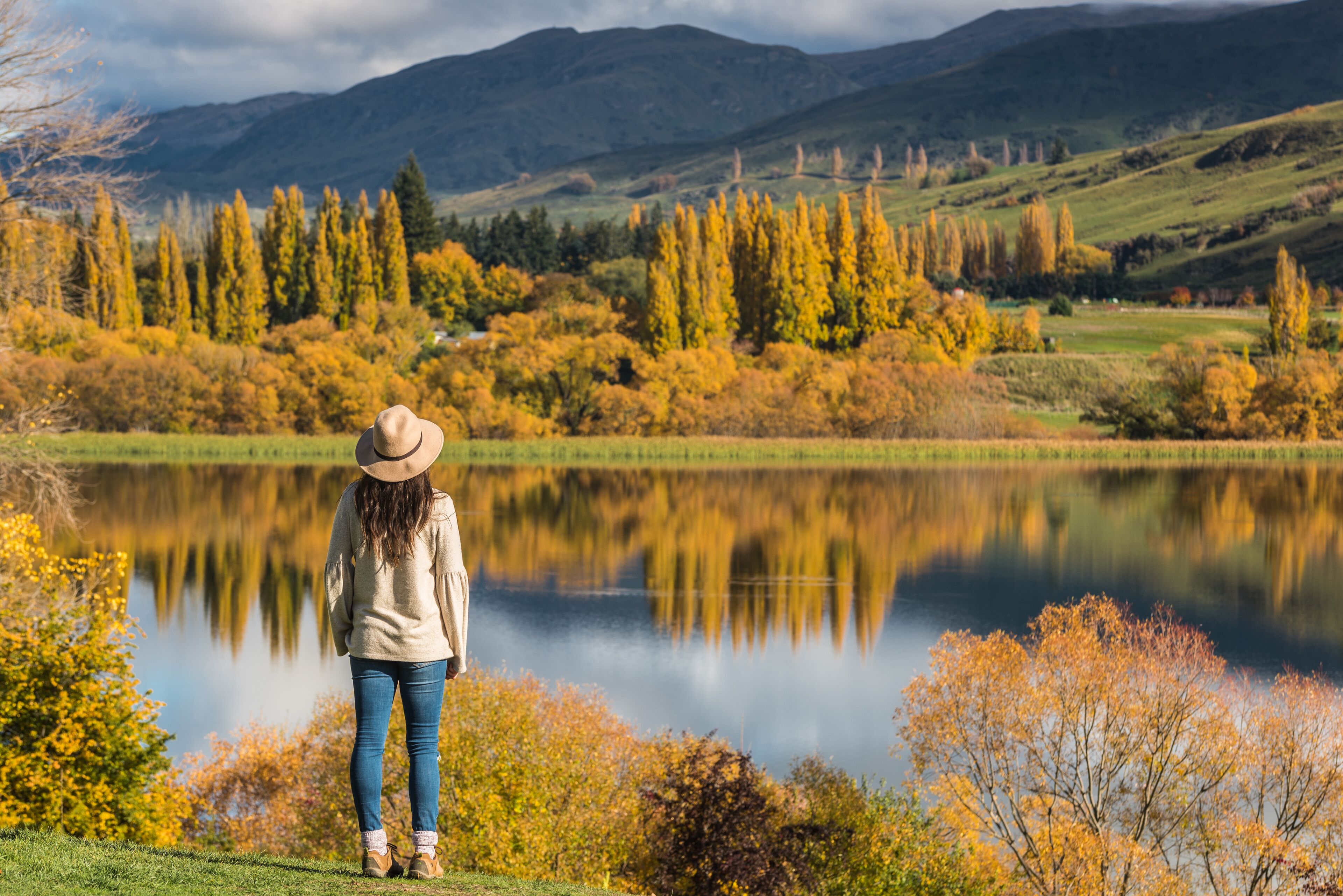 A person standing by a lake with autumn foliage and mountains in the background.