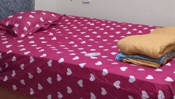 Een eenpersoonsbed met een rood beddegoed met hartjesprint, een wandairconditioner en een grijs gordijn met patroon.