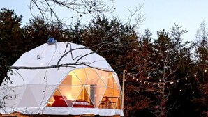 Una tenda a forma di cupola con un pontile di legno, un interno visibile con un letto rosso e luci a stringa sullo sfondo.