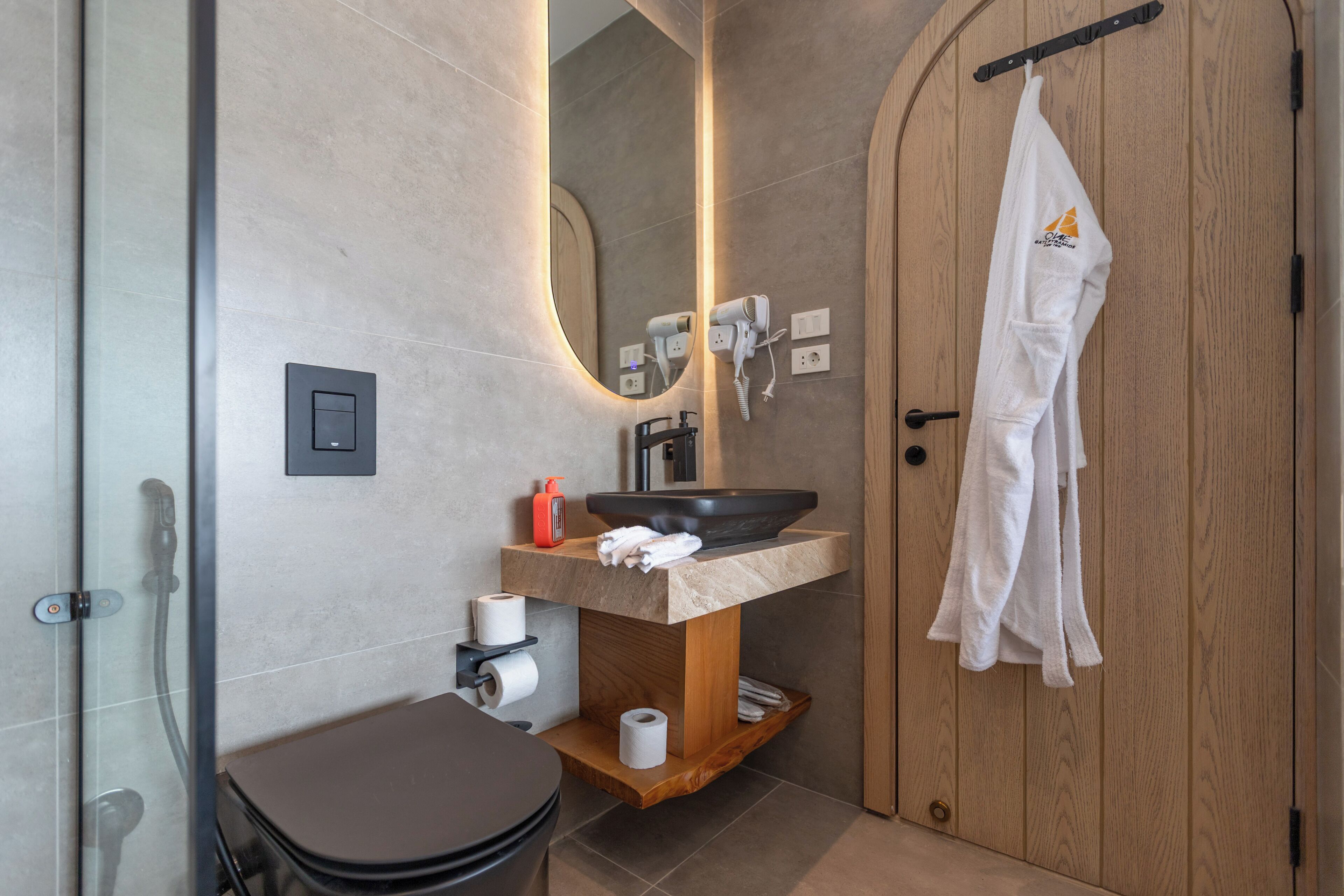 A modern bathroom with a wooden vanity, a large mirror, a black sink, and a towel hanging on the door.