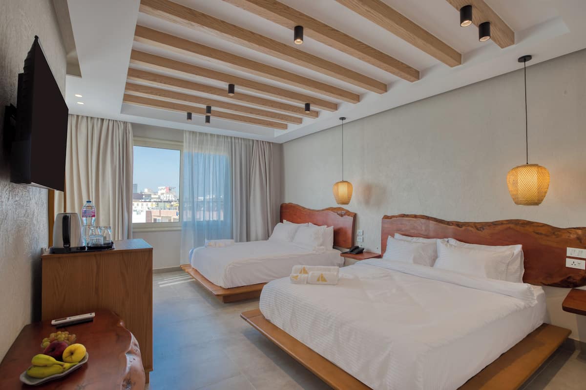 Une chambre d’hôtel avec deux lits, un bureau en bois avec une télévision, un panier de fruits et une vue sur la ville.