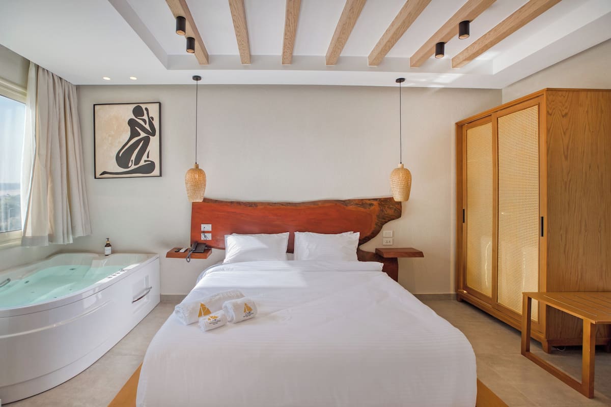 Une chambre d’hôtel avec un grand lit, une tête de lit en bois, une armoire en bois, un banc en bois, une grande fenêtre avec des rideaux, une œuvre d’art accrochée au mur, une table de chevet et une baignoire.