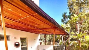 Ein überdachter Patio-Bereich mit Holztisch und Bänken, Steinmauern und Blick auf einen Garten mit Bäumen.