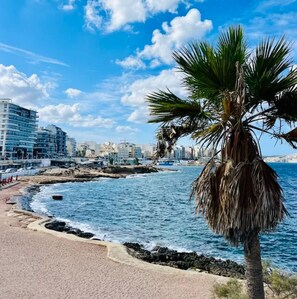 Paesaggio costiero con spiaggia, palme e edifici moderni.