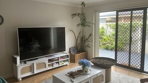 Un salon doté d’un téléviseur à écran plat, d’une table basse avec des livres et un vase, d’une plante, et d’une porte-fenêtre coulissante donnant sur un balcon verdoyant.