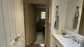 Un couloir étroit, doté d’un plancher en bois, mène à une salle de bain avec un lavabo blanc, un miroir et un porte-serviettes.